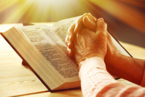 An adult’s praying hands on an open Bible